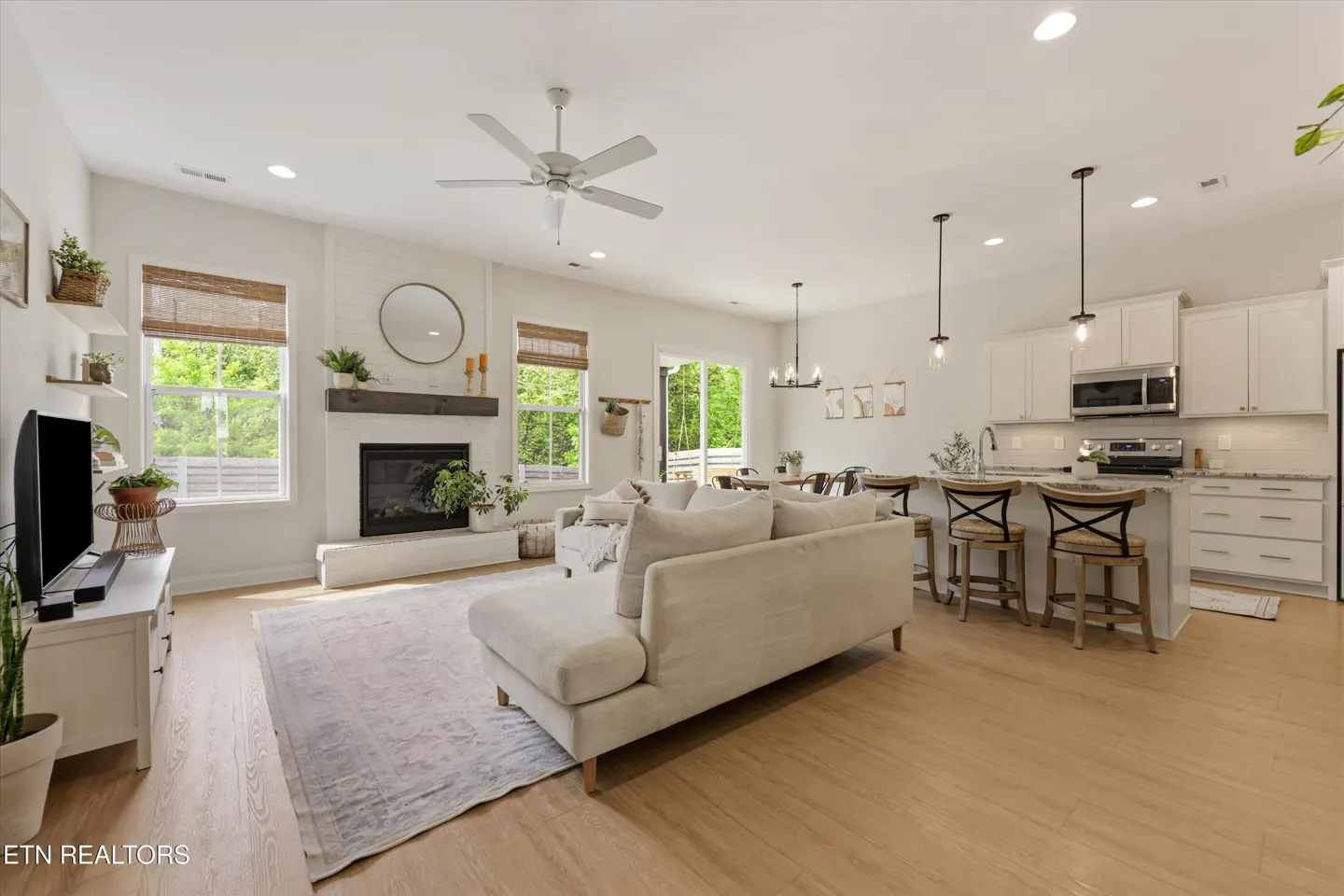 Open-concept living space with a white sofa, fireplace, and kitchen island with bar stools. Light wood floors and natural light.