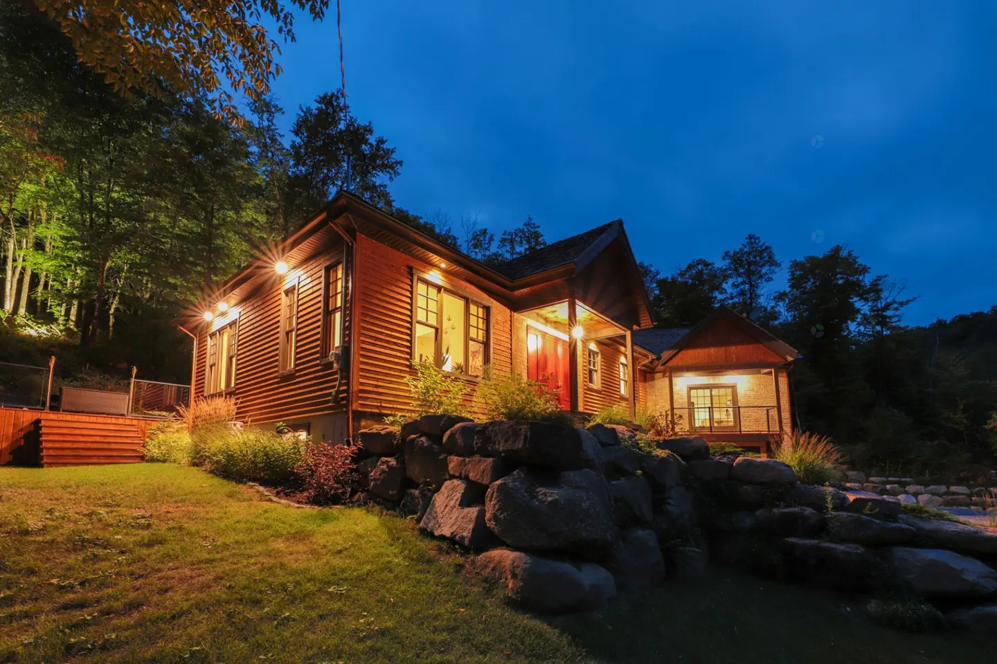 Exterior view of a wood cabin at dusk, with lights on, set on a rocky hill.