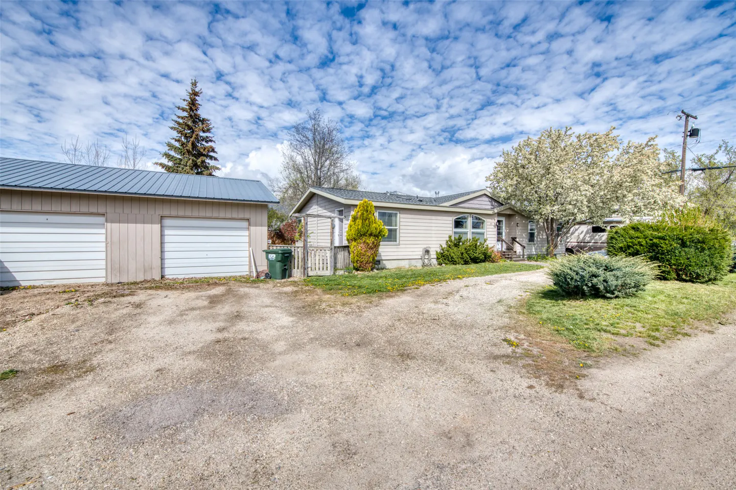 A single-story beige house with a detached two-car garage on a gravel driveway under a cloudy sky.