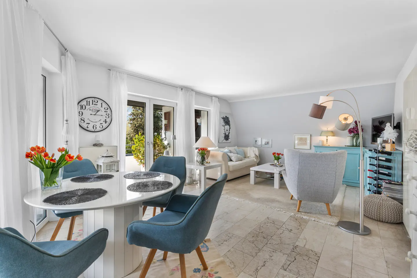 Bright, open-concept living and dining area with a white table, blue chairs, and a gray armchair. A large clock hangs on the wall.