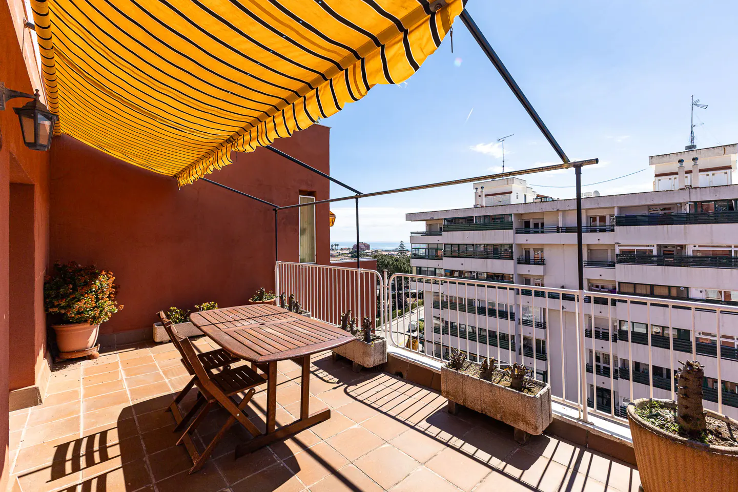 A sunny balcony with terracotta tiles, a wooden table and chairs, and a yellow striped awning. White railings offer city views.