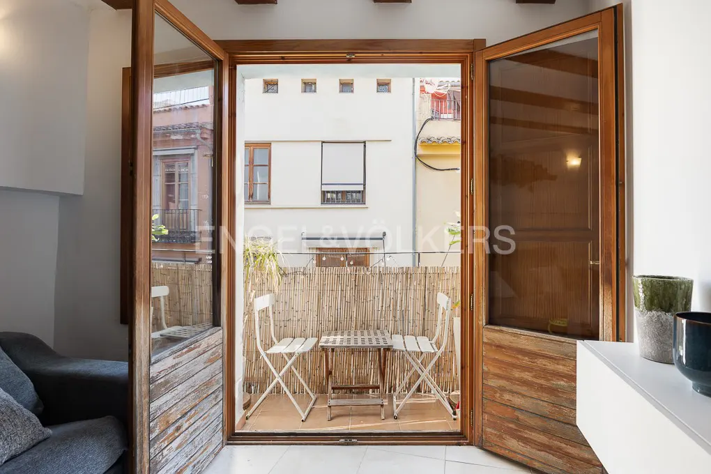 Open wooden doors lead to a balcony with a bamboo screen, two white chairs, and a small table. Buildings are visible in the background.