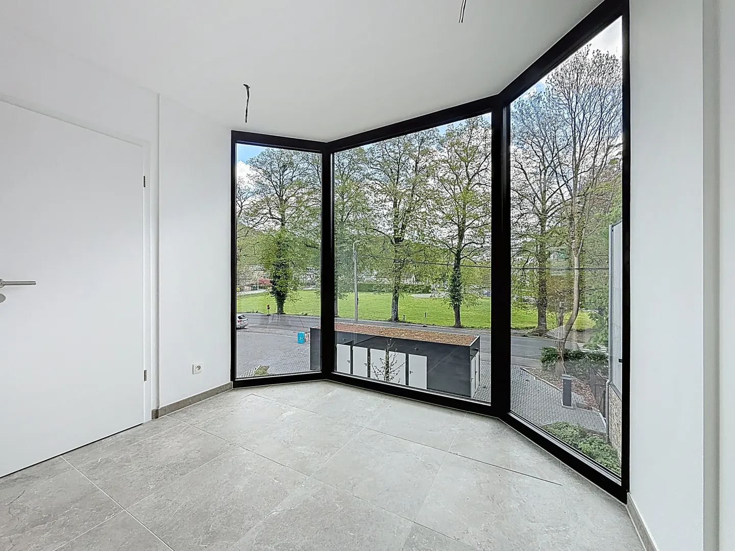 Bright room with gray tile floor, white walls, and a large bay window with black frames overlooking a green park.