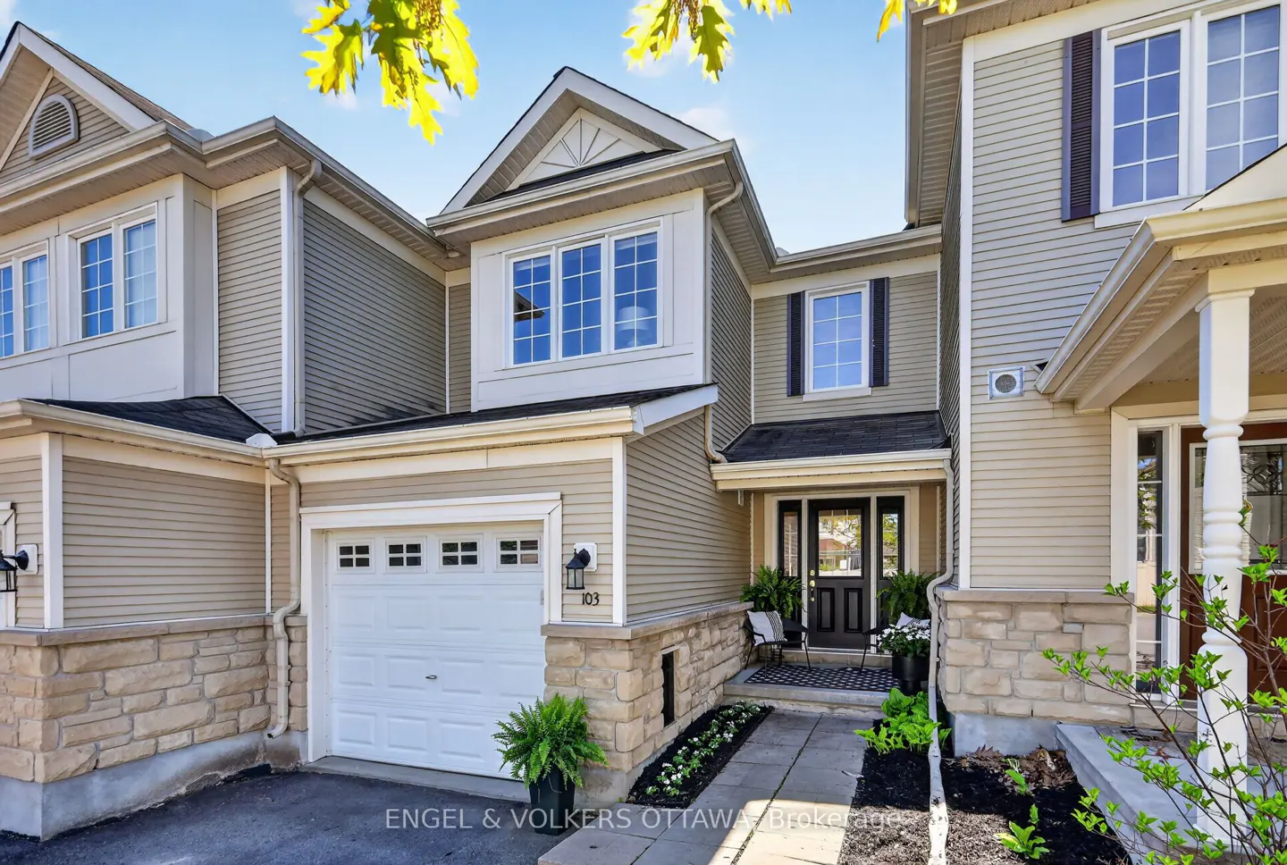 Exterior view of a beige two-story townhouse with a white garage door and black front door. A stone walkway leads to the entrance.