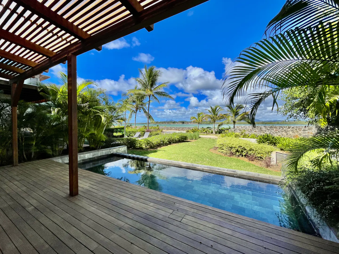 View from a wooden deck with a pergola, overlooking a pool, lawn, palm trees, and ocean under a blue sky with clouds.