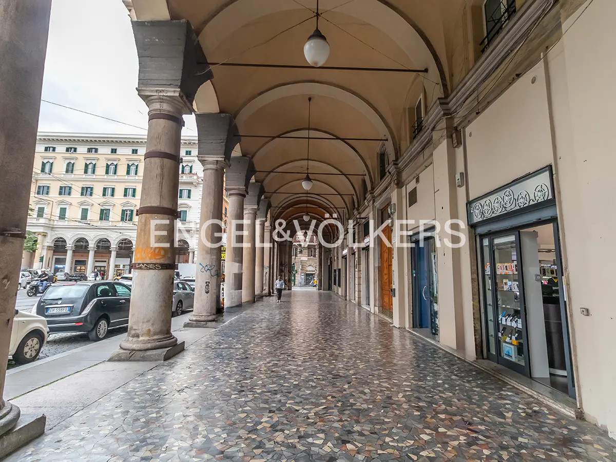 Covered walkway with stone columns and arches, leading to shops. Cars parked on the street. The floor is mosaic tile.