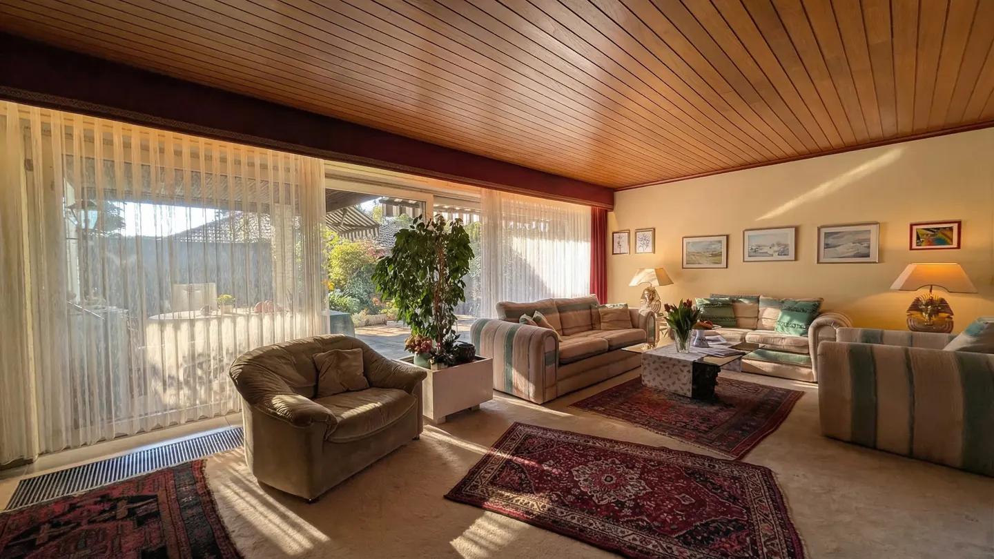 Living room with wood ceiling, striped sofas, and red rugs. Sunlight streams through sheer curtains. A potted plant sits near a sliding glass door.