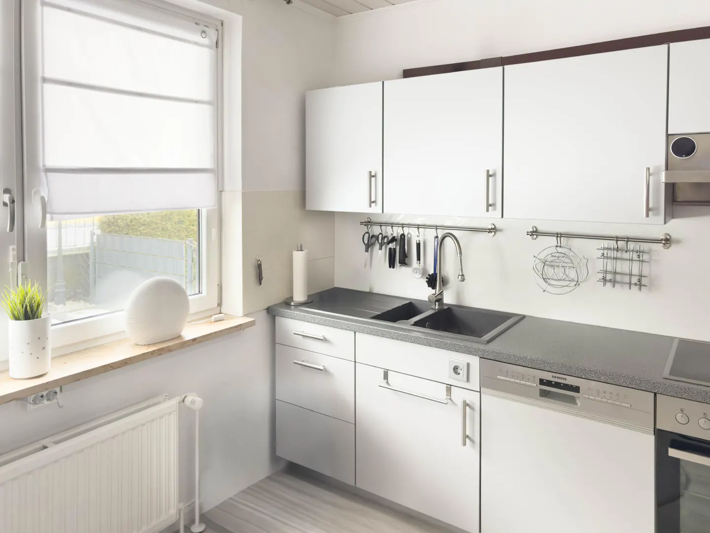 Bright kitchen with white cabinets, gray countertops, and a black sink. Utensils hang above the sink. A window with a white shade is to the left.
