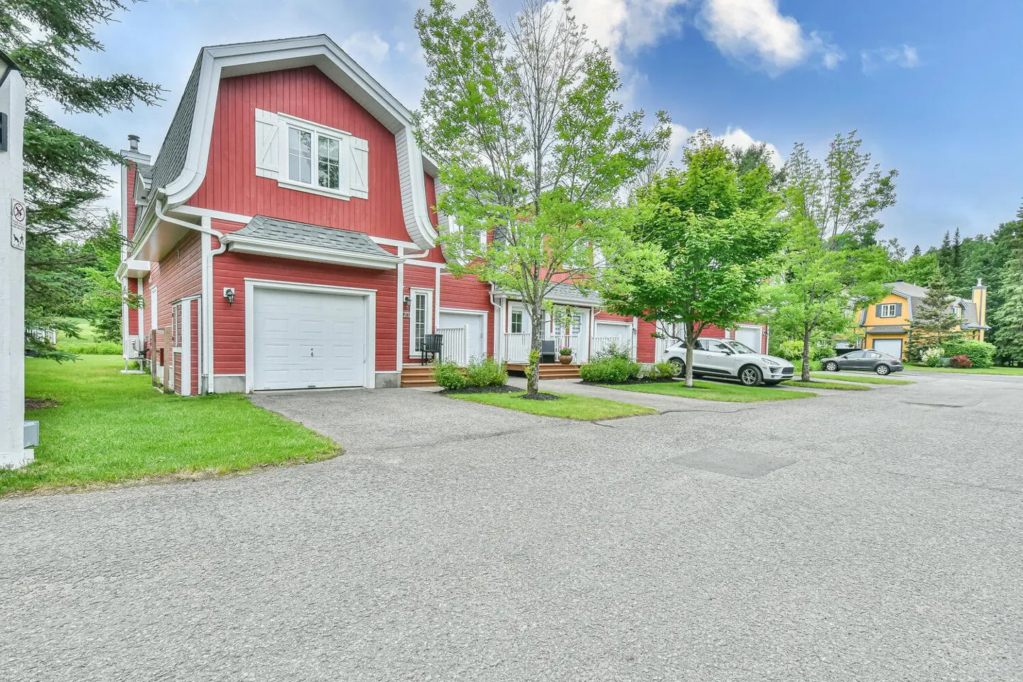 Red townhouses with white trim and garage doors line a paved driveway. Green trees and grass surround the buildings under a blue sky.
