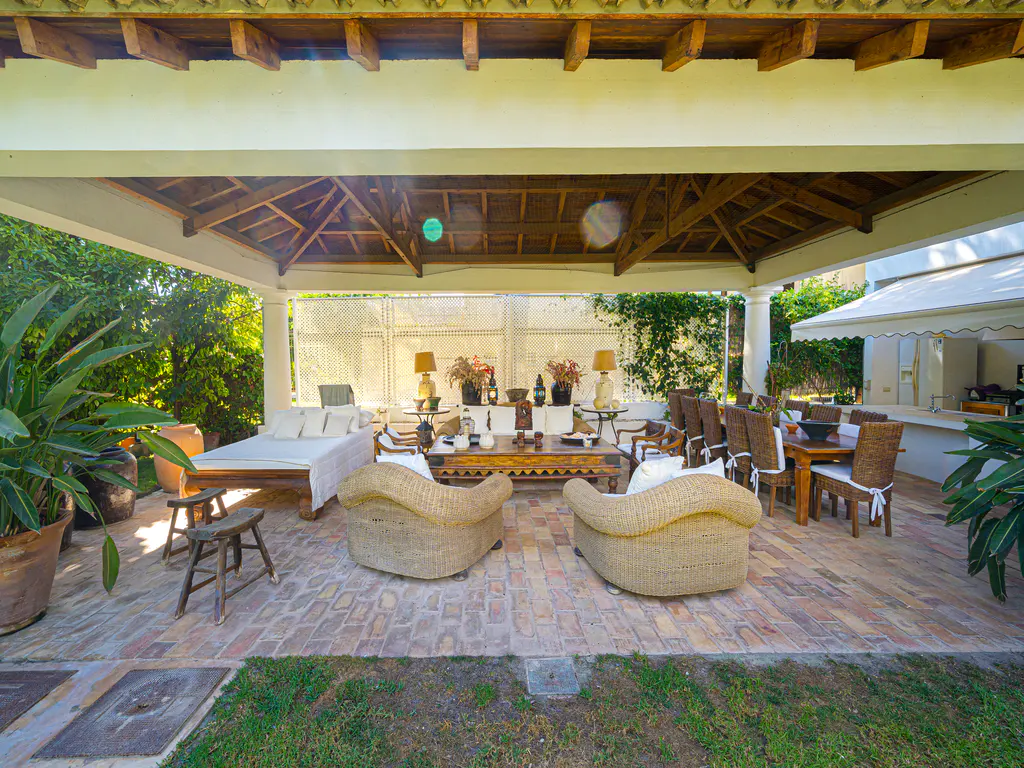 Covered outdoor patio with brick flooring, wicker armchairs, a wooden coffee table, and a dining area with wicker chairs.