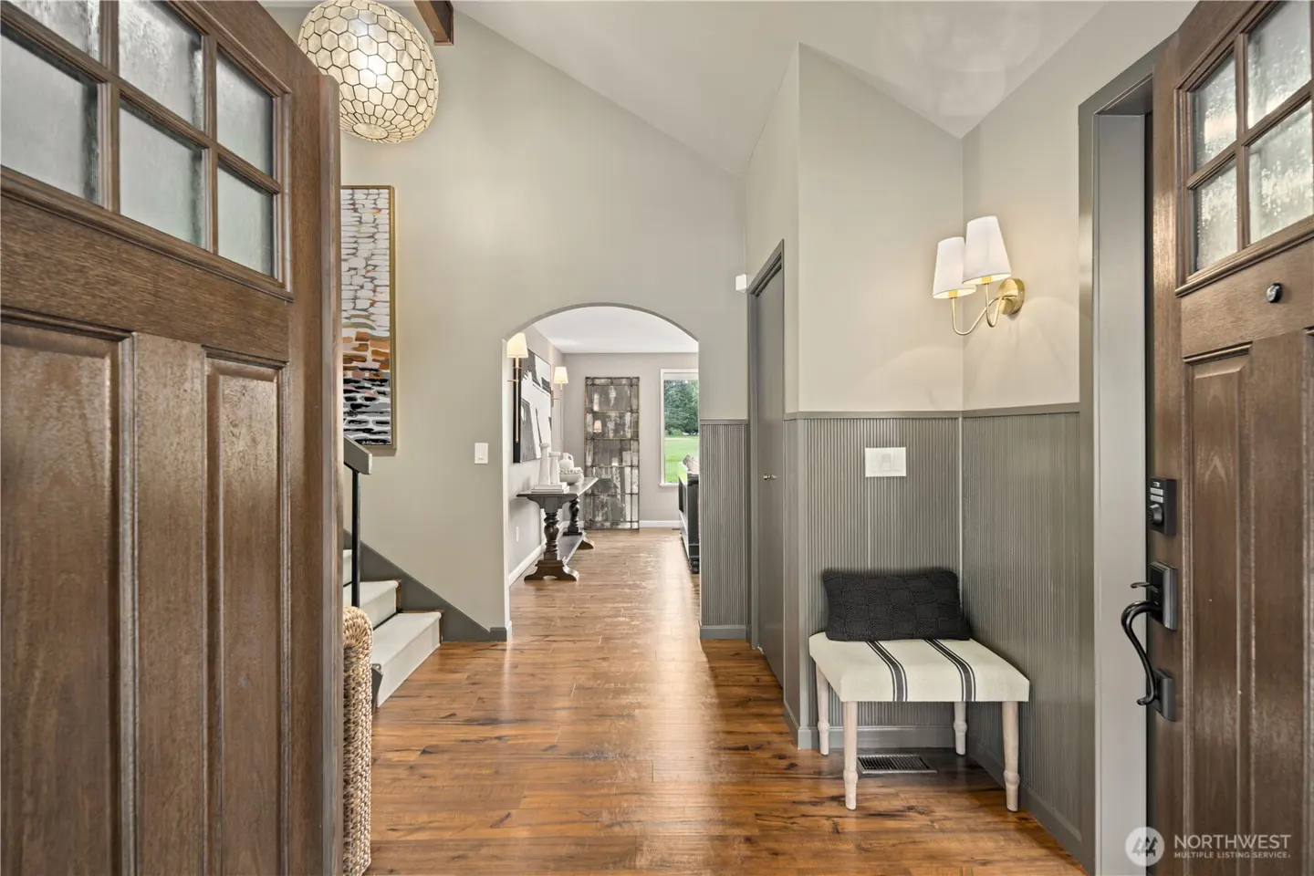 Open front door reveals a foyer with hardwood floors, gray walls, and a bench. A hallway leads to a view of the outdoors.
