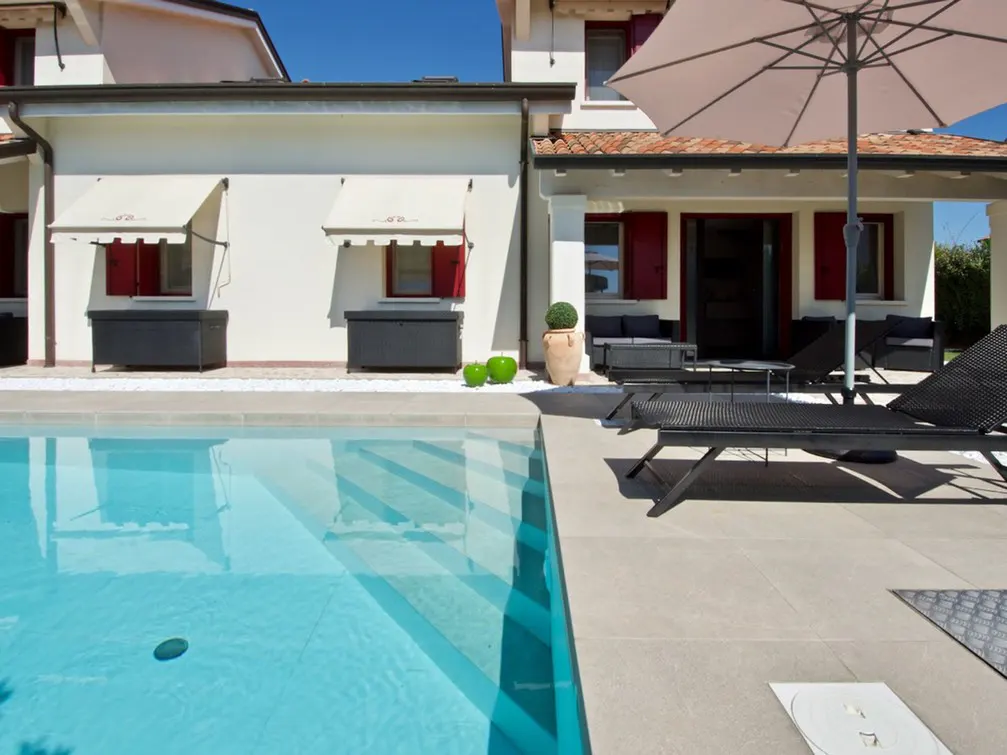 A bright, sunny backyard with a pool, lounge chairs, and a white house with red shutters.