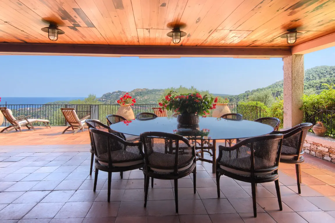 Covered patio with a round table, chairs, and ocean view. The patio has a wood ceiling, tile floor, and potted plants.