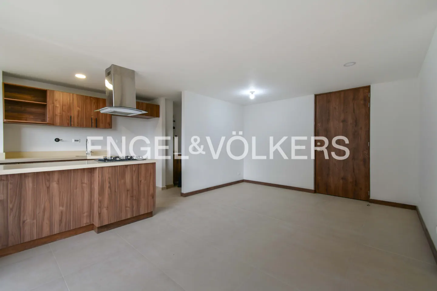 Modern kitchen with wood cabinets, stainless steel range hood, and a matching island, opening into a spacious white room.