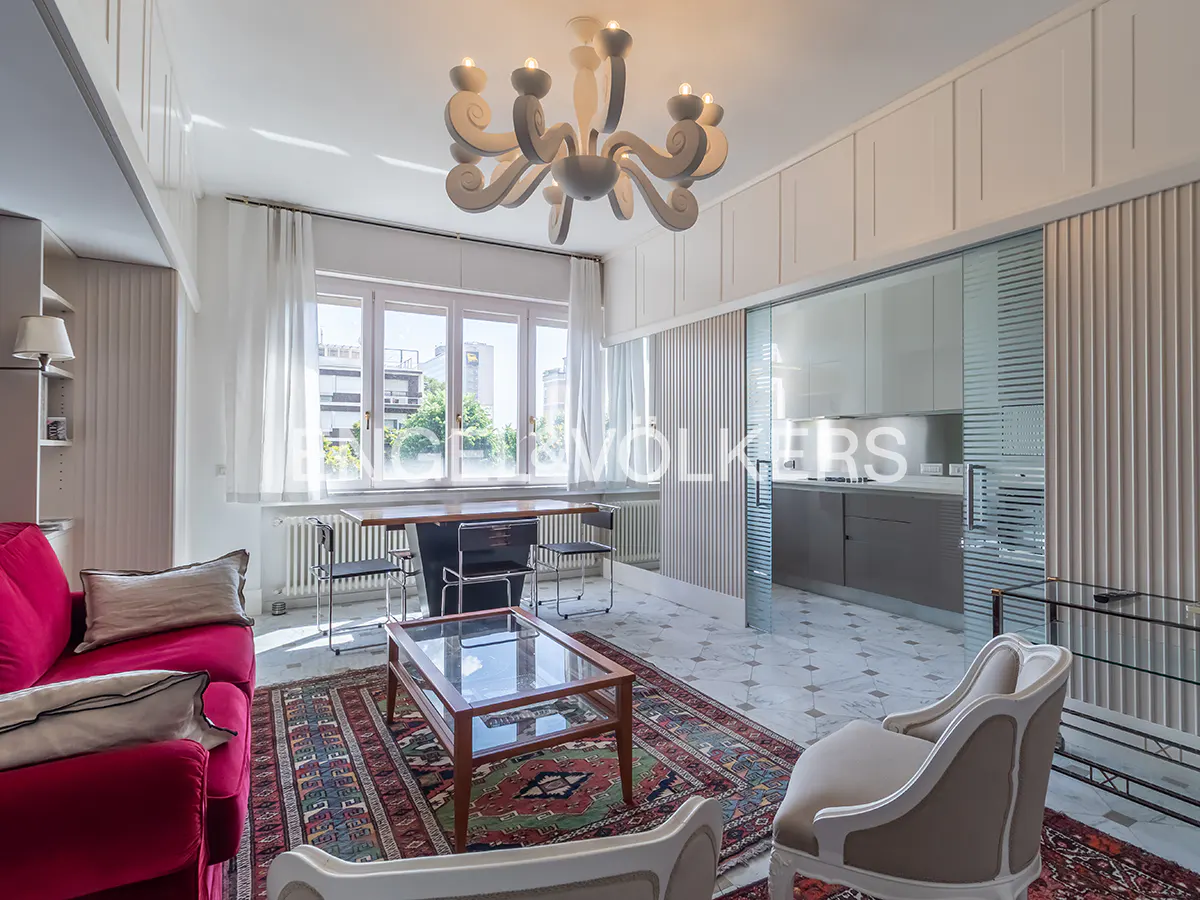 Bright living room with red sofa, patterned rug, glass table, and white chairs. A modern chandelier hangs above. Kitchen visible through glass doors.