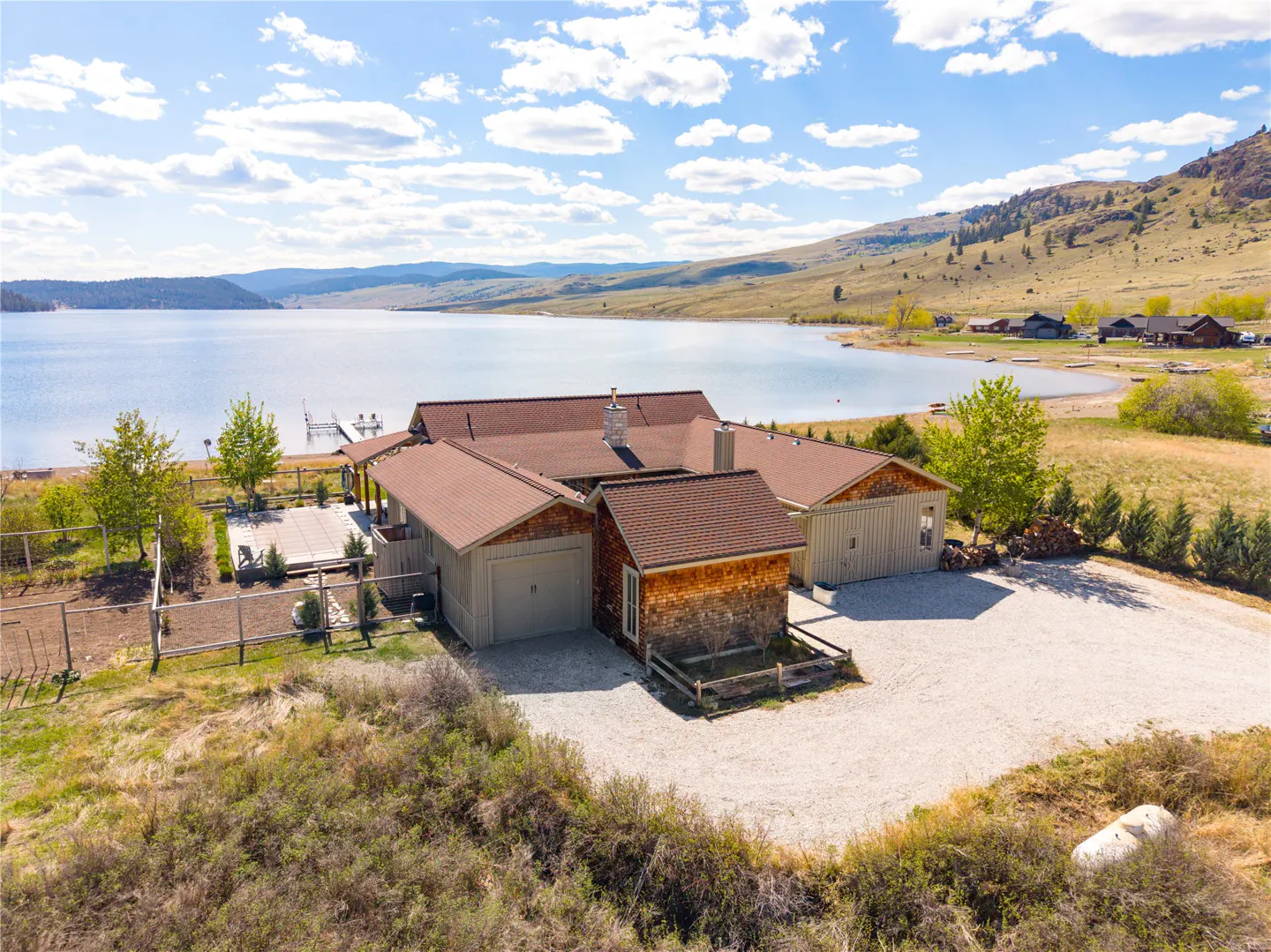 Lakefront home with a brown roof, beige siding, and a gravel driveway. A lake and mountains are in the background under a blue sky with clouds.