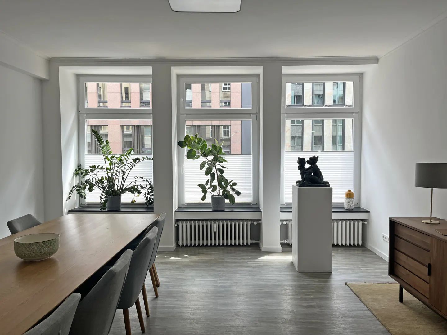 Bright room with three windows, plants, and a sculpture. A wooden table and gray chairs are on the left. A wooden cabinet is on the right.