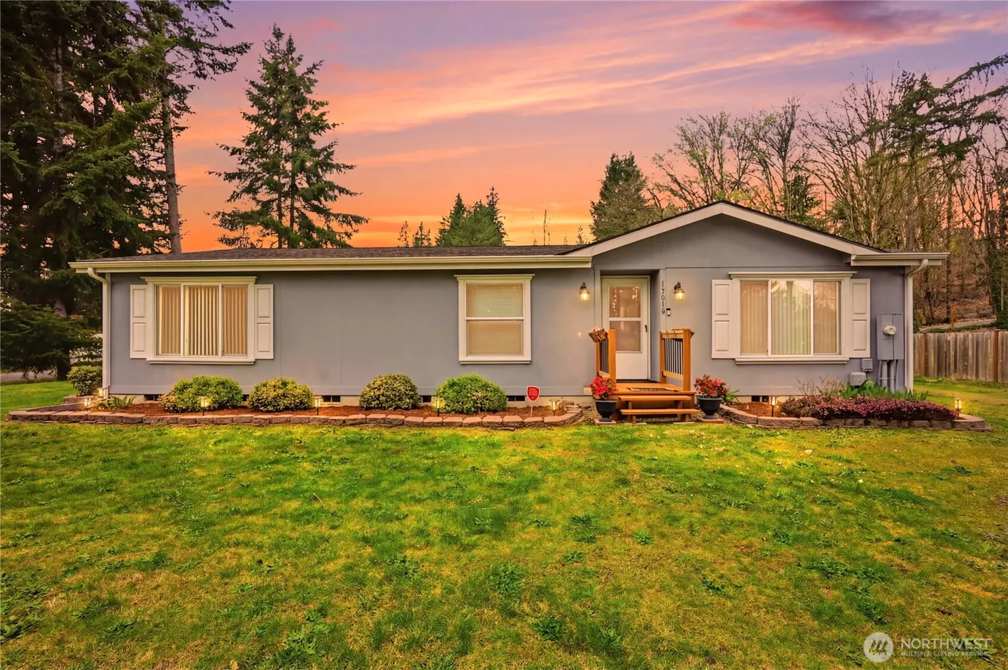 A single-story gray house with white shutters, a small porch, and a green lawn under a colorful sunset sky.