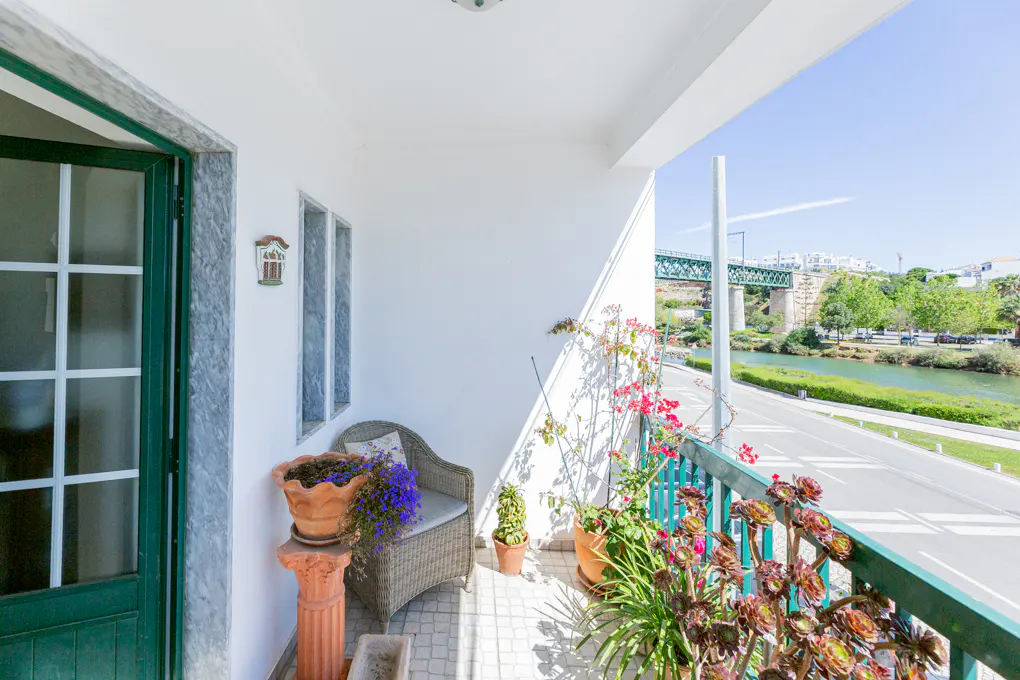 Balcony view with green railing, potted plants, and wicker chair. A bridge and river are visible in the background.