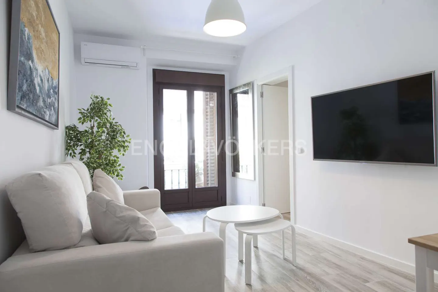 Bright living room with white walls, light wood floors, and a white sofa. A large TV hangs on the wall, and a balcony door lets in natural light.