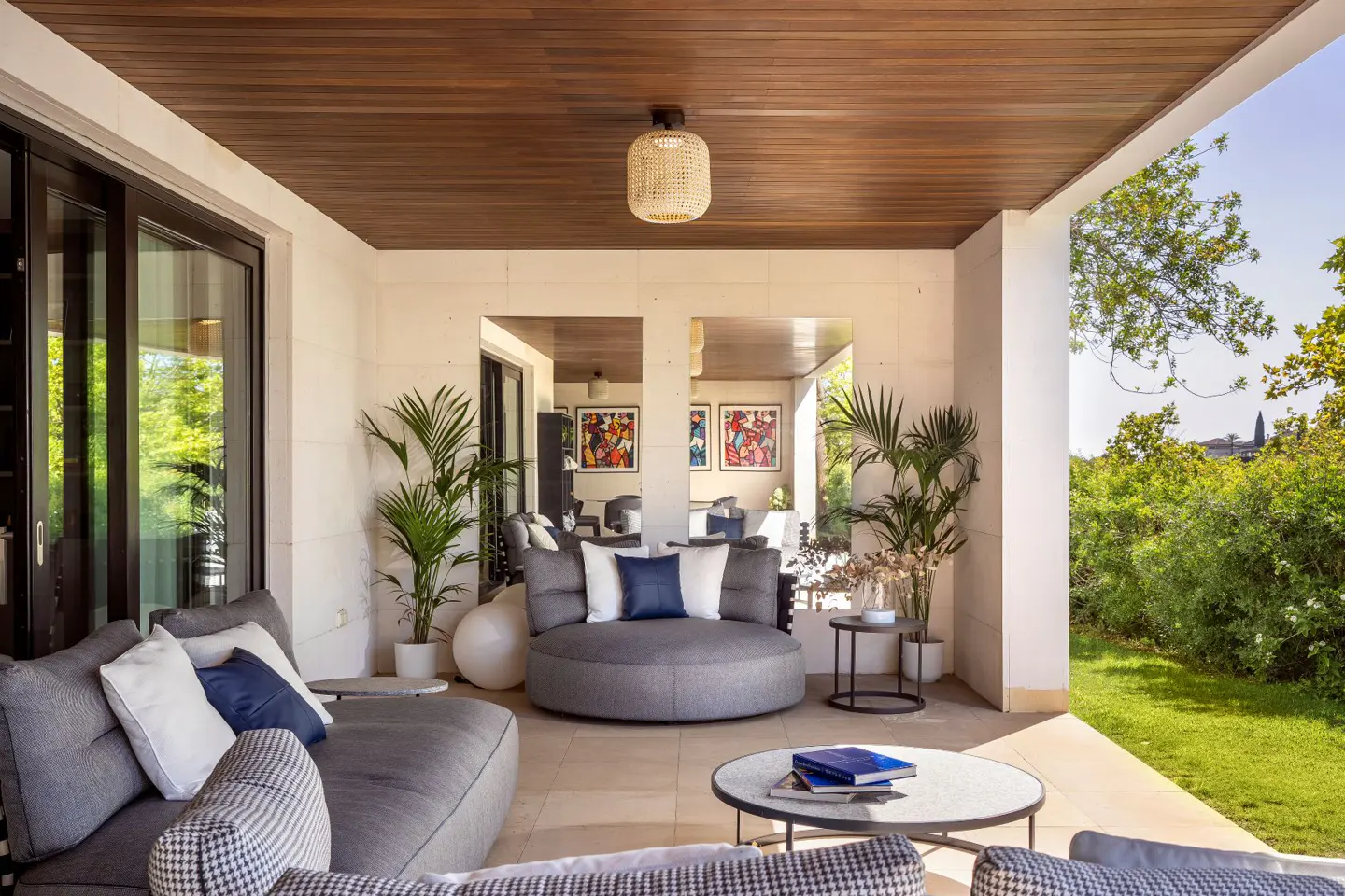 Covered patio with gray couches, blue pillows, and a round coffee table with books. Green trees are visible in the background.