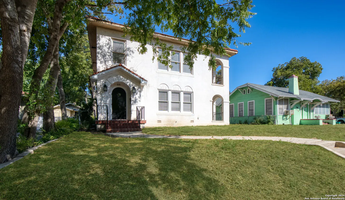Two-story white stucco home with a front porch and arched entryway, next to a light green house under a clear blue sky.