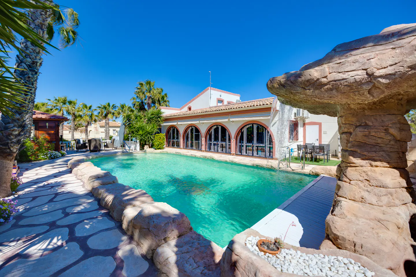 Outdoor pool with turquoise water, rock features, and a white building with arched windows under a clear blue sky. Palm trees surround the area.