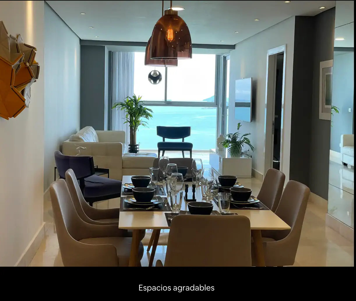 Modern apartment dining area with table set for eight, overlooking ocean view. Pendant lights hang above.