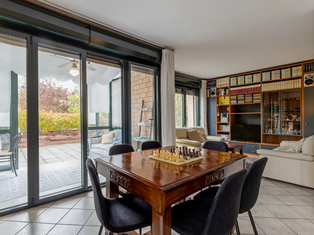 Interior view of a dining room with a chess board on a wooden table and black chairs. Sliding glass doors lead to a patio. A bookcase is in the background.