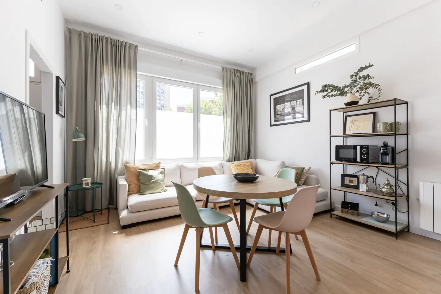 Bright living room with a white sofa, round table with pastel chairs, and a black metal shelving unit against a white wall.