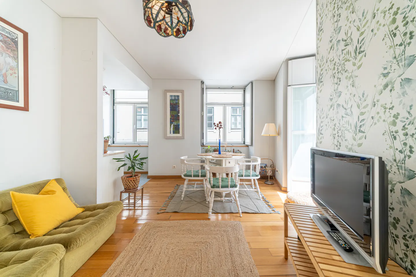 A bright living room with hardwood floors, a green couch with a yellow pillow, and a white dining set. A TV sits on a wooden stand against a leafy wallpaper.