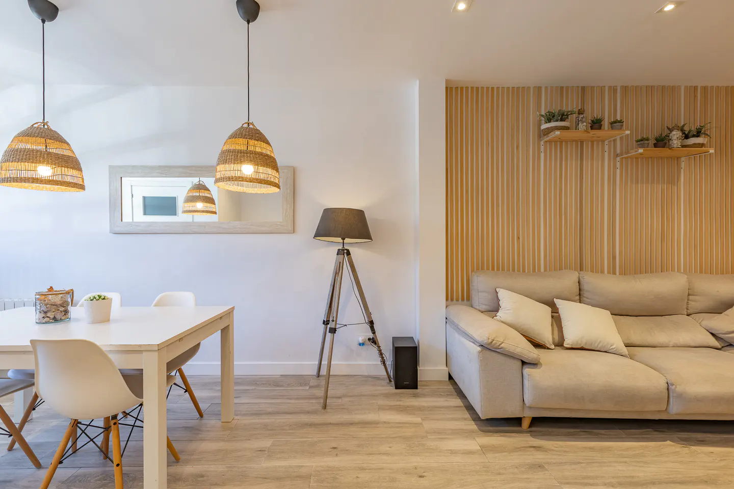 Living room with a white dining table, beige sofa, wood-paneled wall, and wicker pendant lights. A tripod floor lamp stands near the sofa.