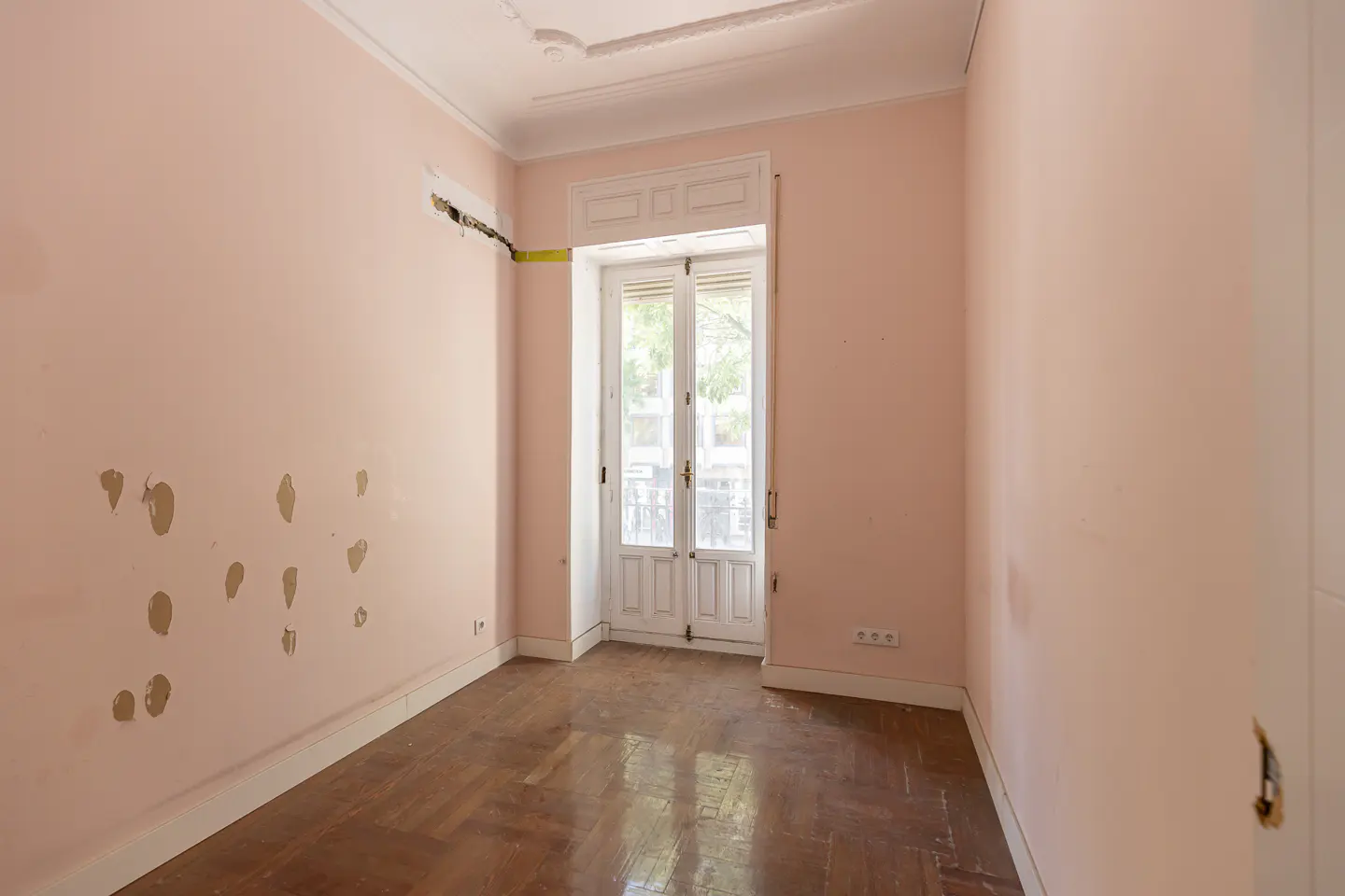 Empty room with pink walls, wooden parquet flooring, and a white balcony door leading to an outdoor view.