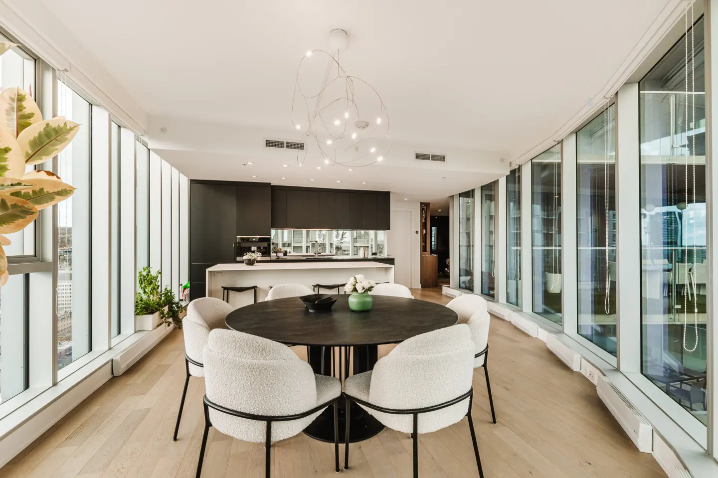 Bright, modern condo interior with floor-to-ceiling windows. A round, dark wood table is surrounded by white chairs. Black cabinets and a white island define the kitchen.