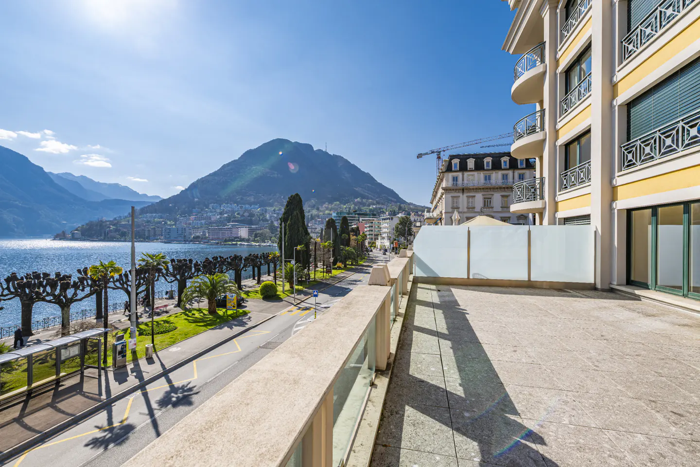 View from a balcony overlooking a lakeside road, with mountains in the background under a clear blue sky.
