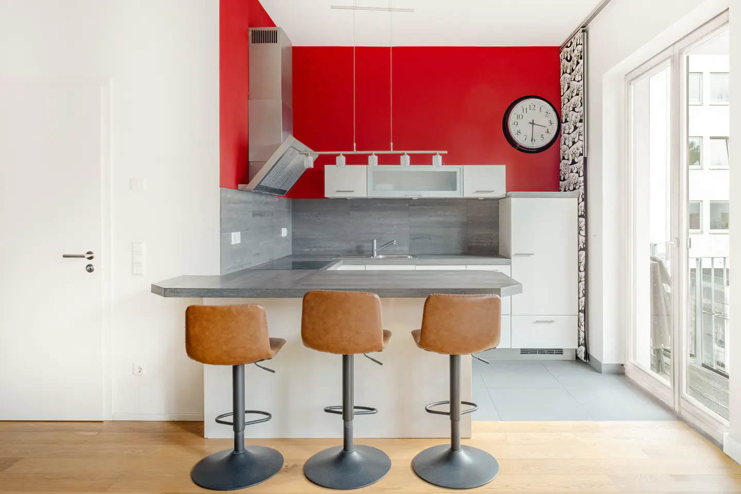 A modern kitchen with a red accent wall, stainless steel appliances, and three brown leather bar stools.