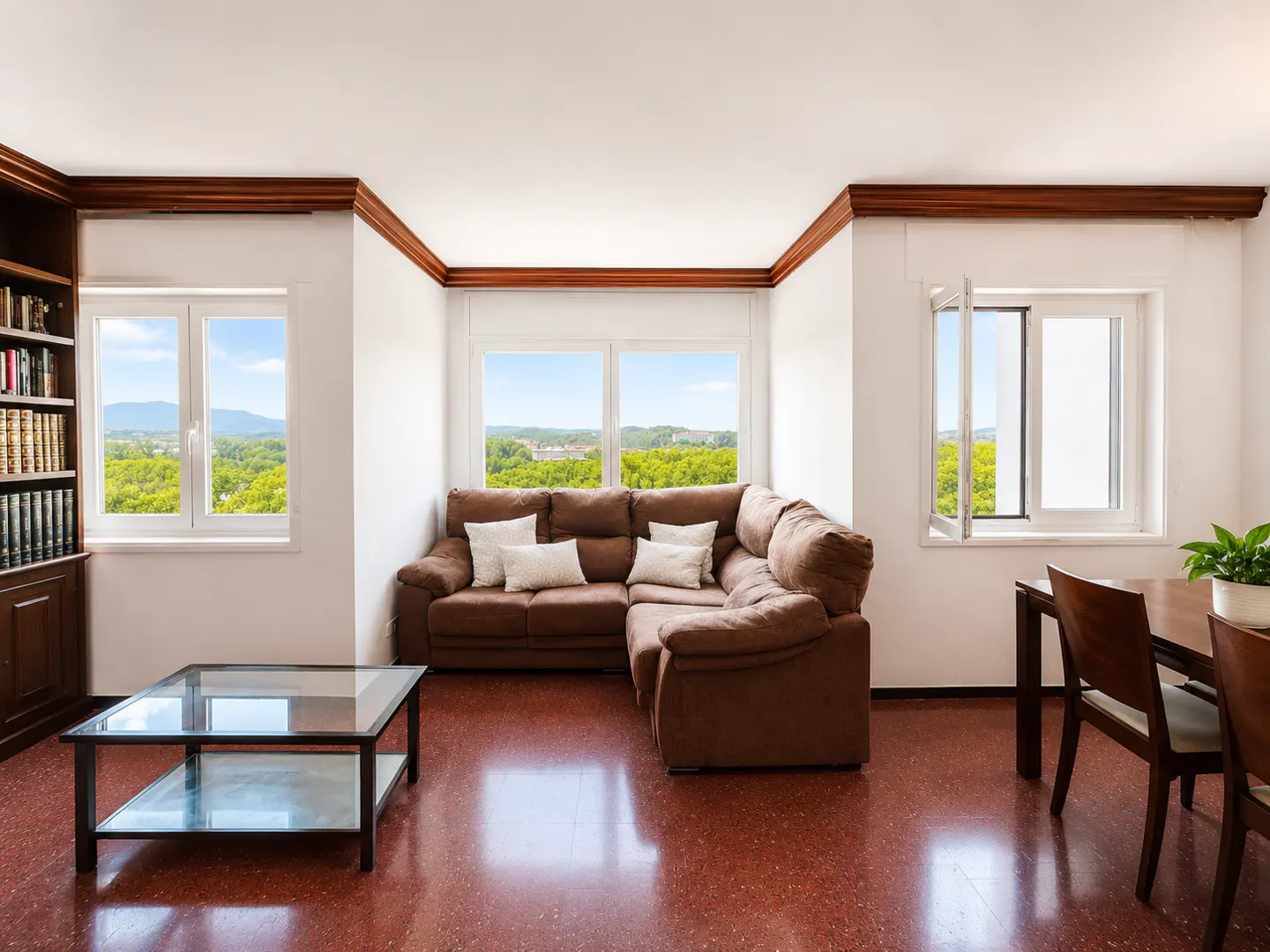 Living room with a brown sectional sofa, glass coffee table, wooden bookshelf, and dining area overlooking a green landscape.