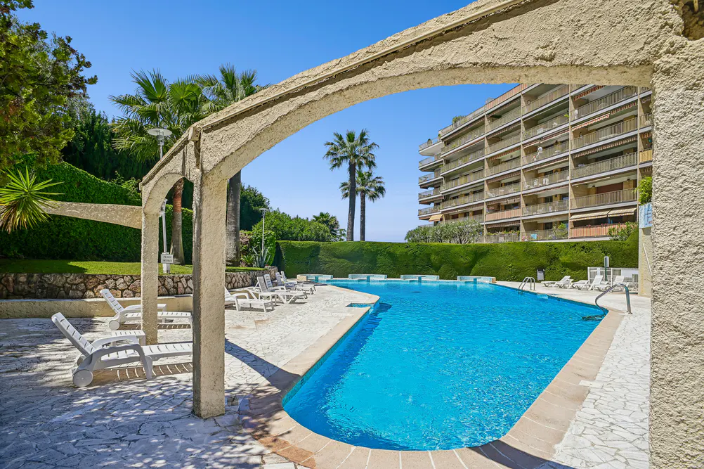 Outdoor pool with lounge chairs, viewed through a stone archway. Palm trees and a multi-story building are in the background.