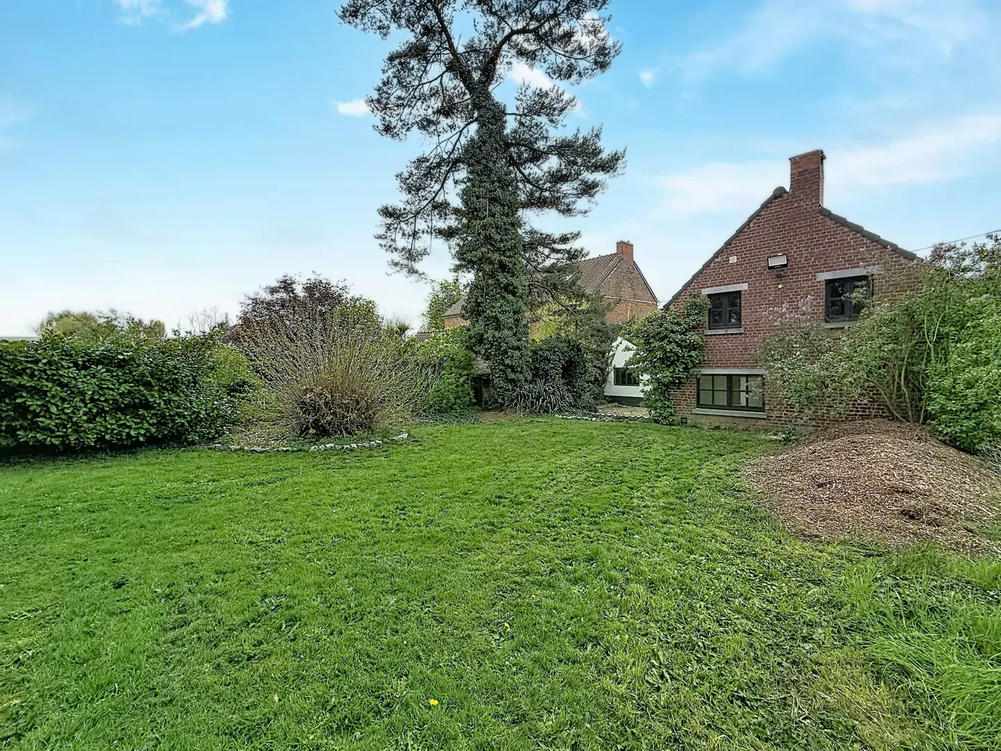A brick house with black windows sits behind a green lawn and trees under a blue sky.