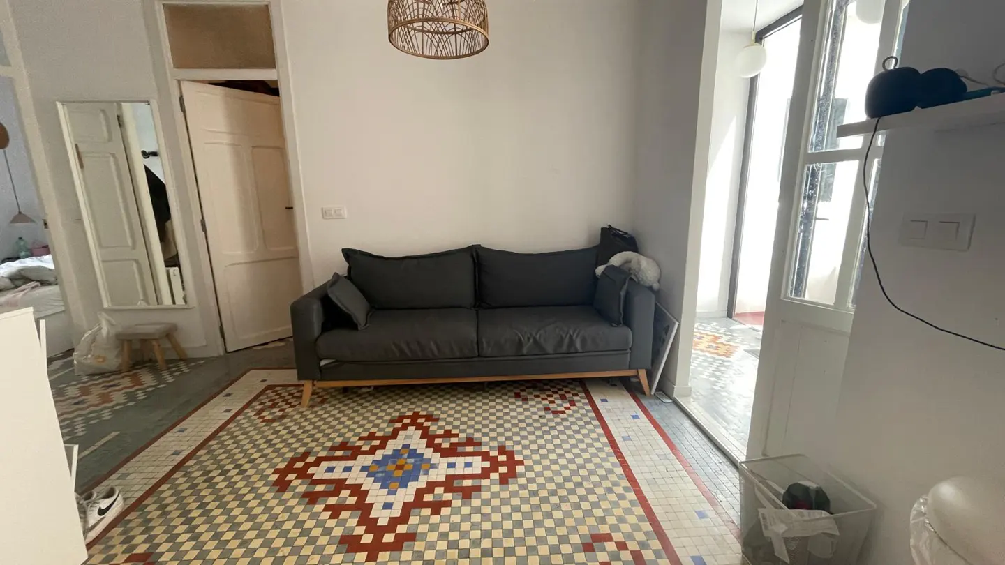 Living room with a gray sofa on a patterned tile floor. A doorway leads to another room with a bed. Natural light streams in from a window.
