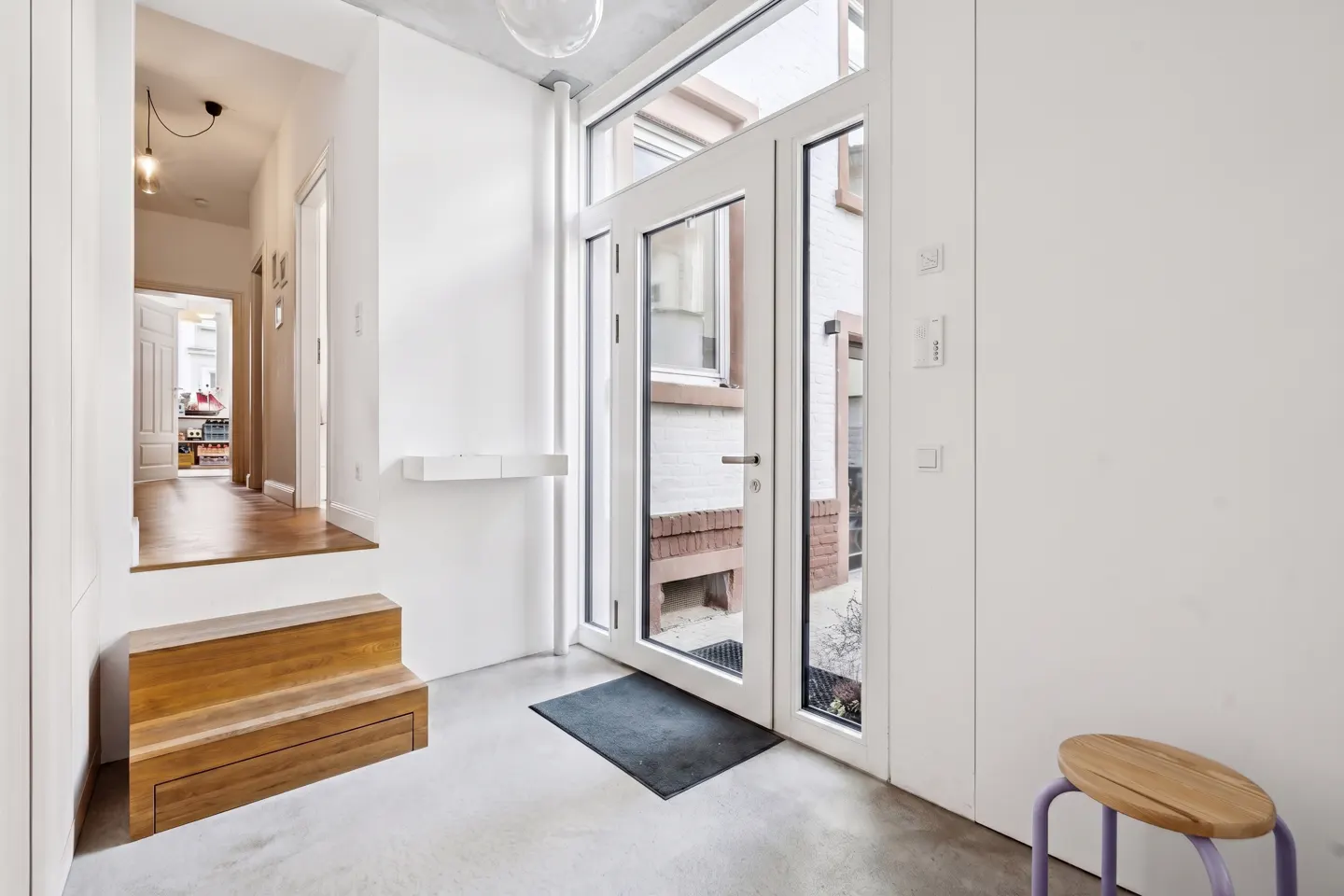 Bright entryway with white walls, a wooden staircase, and a glass-paneled door leading outside. A small wooden stool sits near the door.