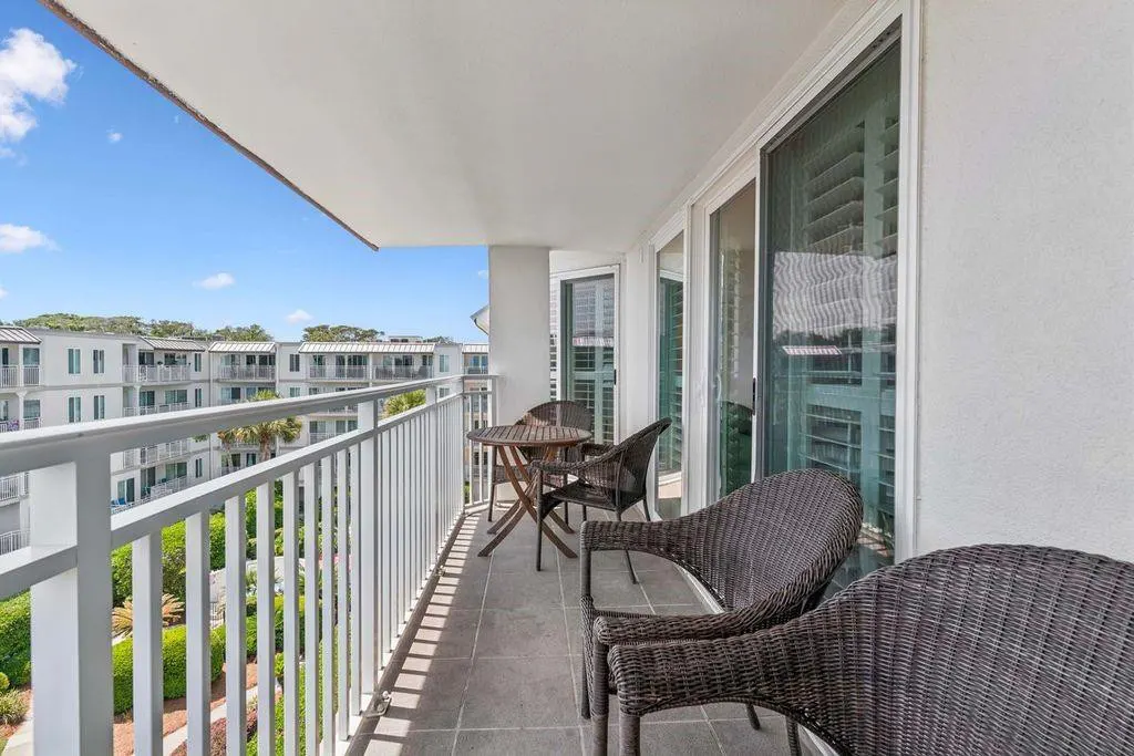 Balcony with wicker chairs and a small table overlooks a green landscape and white buildings under a blue sky.