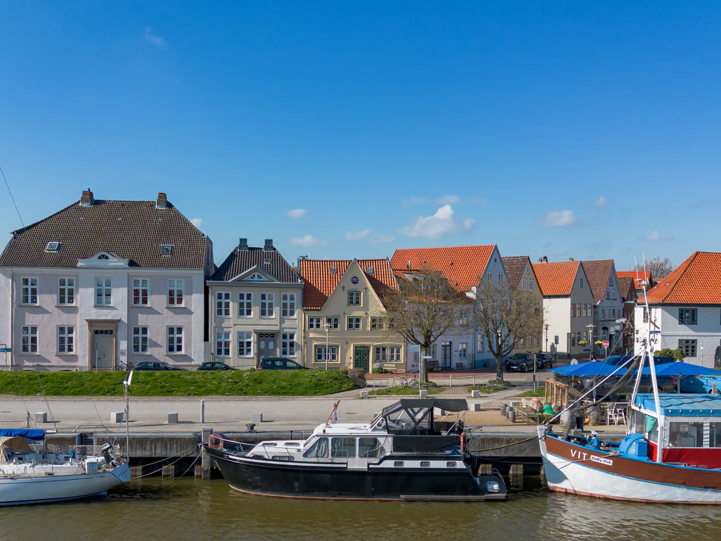 Waterfront view of colorful houses and boats under a blue sky. Boats are docked along a concrete pier.