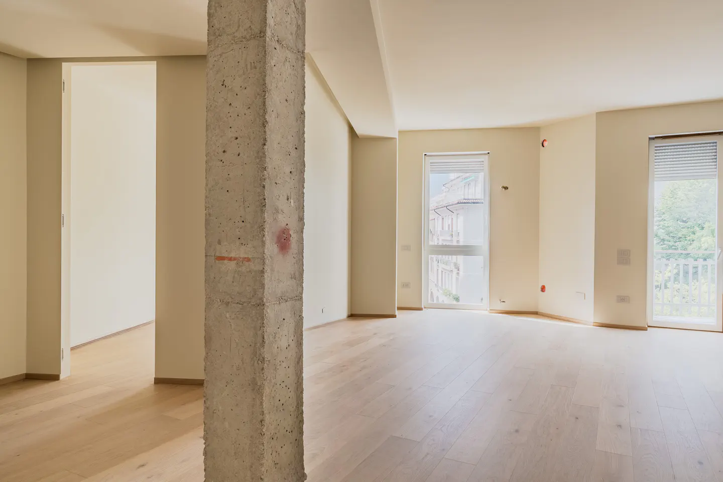 Empty room with light wood floors, beige walls, a raw concrete pillar, and two windows overlooking a city street.