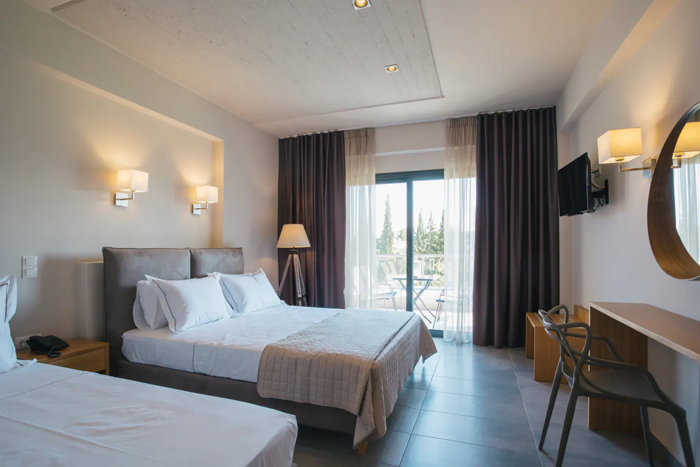 Modern hotel room with a double bed, white linens, dark curtains, a balcony, and a desk with a chair.