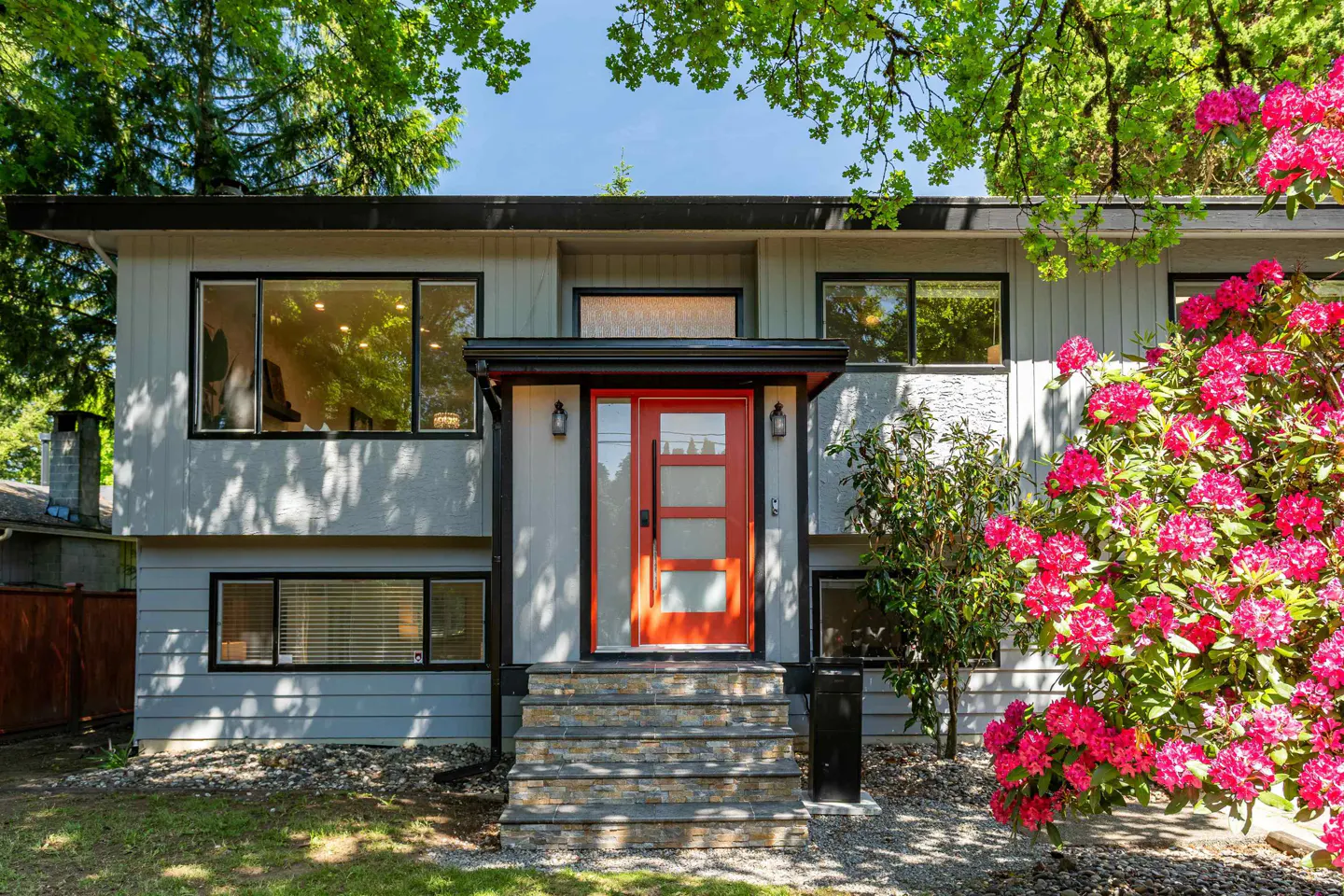 Two-story gray house with a bright orange front door and stone steps. Pink rhododendrons bloom on the right.