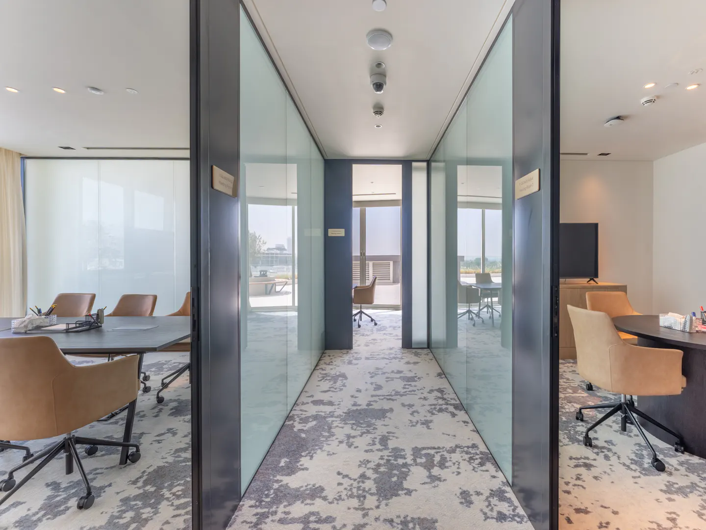 Office interior with glass-walled rooms, a patterned carpet, and conference tables with tan chairs.