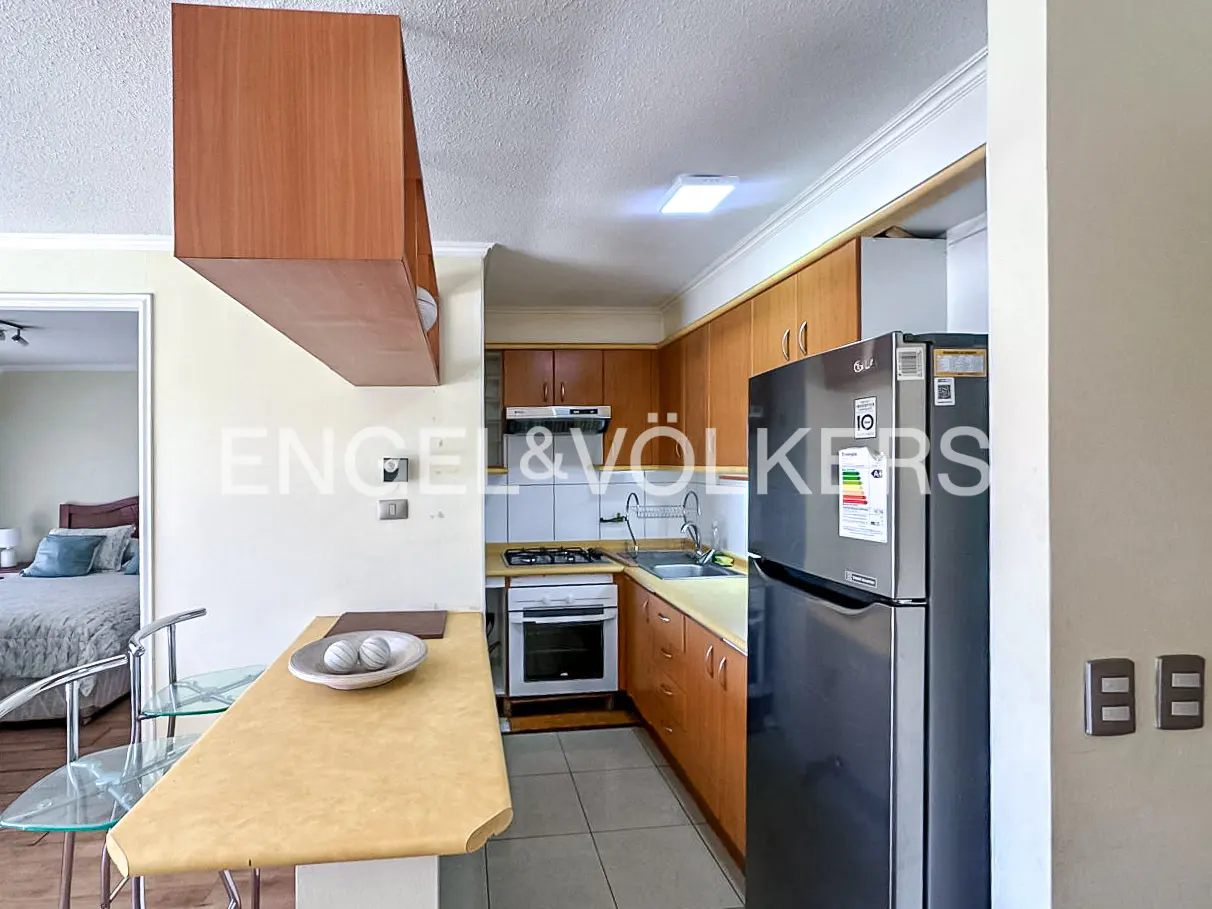 A kitchen with wood cabinets, a gray refrigerator, and a breakfast bar with a glass chair. A bedroom is visible through a doorway.