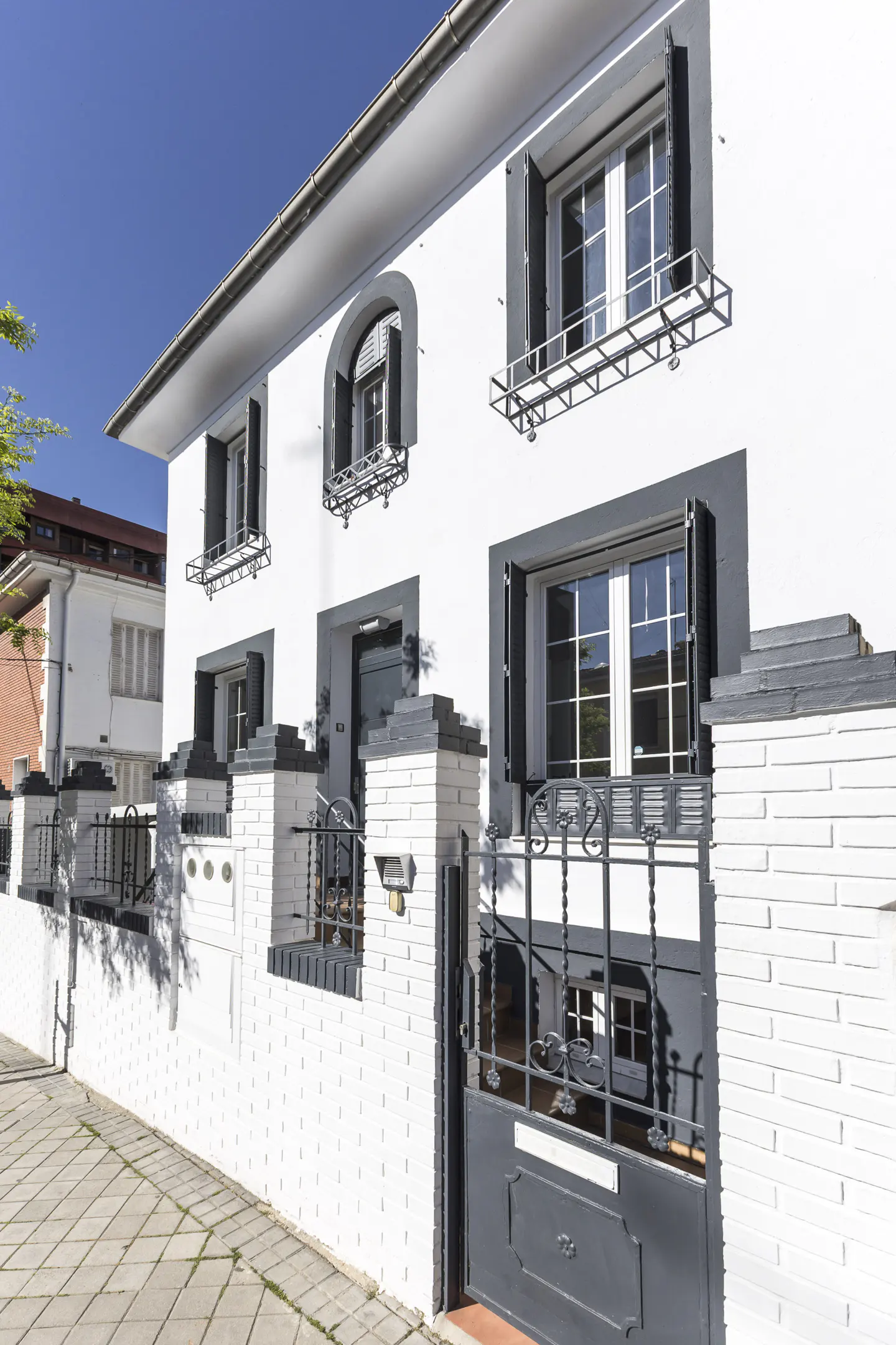 Two-story white house with black shutters and a gray gate. The house has a white brick fence and a blue sky in the background.