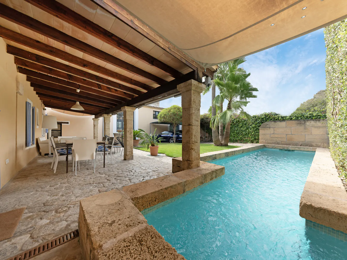 Outdoor patio with stone flooring, wooden beams, and a turquoise pool surrounded by greenery.
